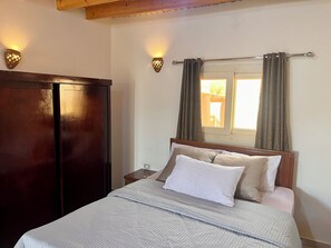 Ein Schlafzimmer mit einem Bett, einem Kleiderschrank, zwei Wandleuchten, einem Fenster mit Vorhängen und einer Holzbalkendecke.