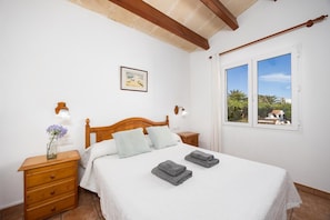 Un dormitorio con cama de madera, ropa de cama blanca, una mesita de noche con un florero, una ventana con vistas y un cuadro en la pared.