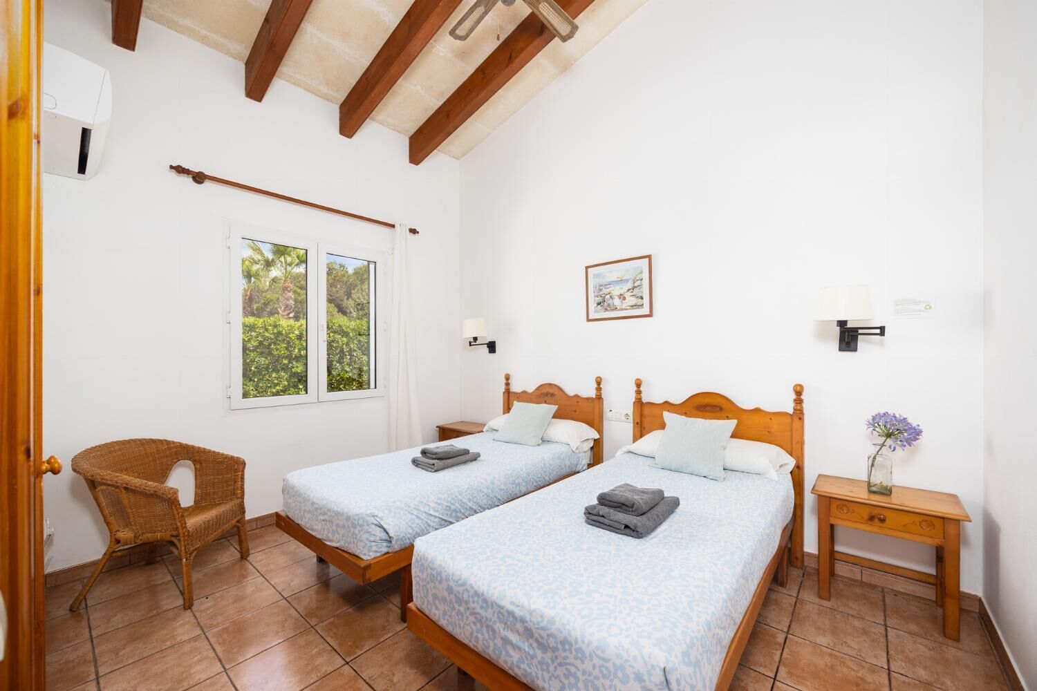 Une chambre avec deux lits, une chaise en osier, une fenêtre donnant sur un beau paysage et une table de chevet avec une lampe.