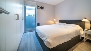 Une chambre moderne avec un grand lit, une table de chevet, une lampe fixée au mur et une porte-fenêtre coulissante donnant sur un balcon.