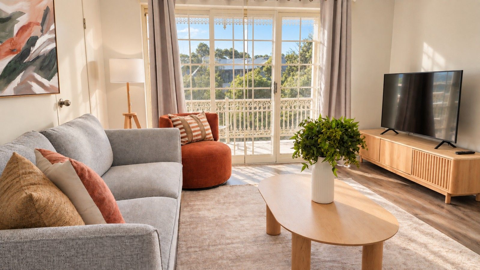 A living room with a grey sofa, a red armchair, a wooden coffee table, a TV on a wooden stand, and a view of greenery outside.
