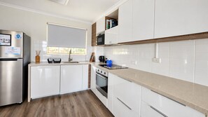 A modern kitchen with white cabinetry, a stainless steel refrigerator, a built-in oven, and a window with a white blind. - 2BR Inner South Stay with Pools, Gym and Sauna (Griffith)
