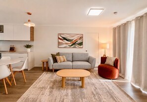 A modern living room with a sofa, a red armchair, a coffee table, and a dining area with chairs. - 2BR Inner South Stay with Pools, Gym and Sauna (Griffith)