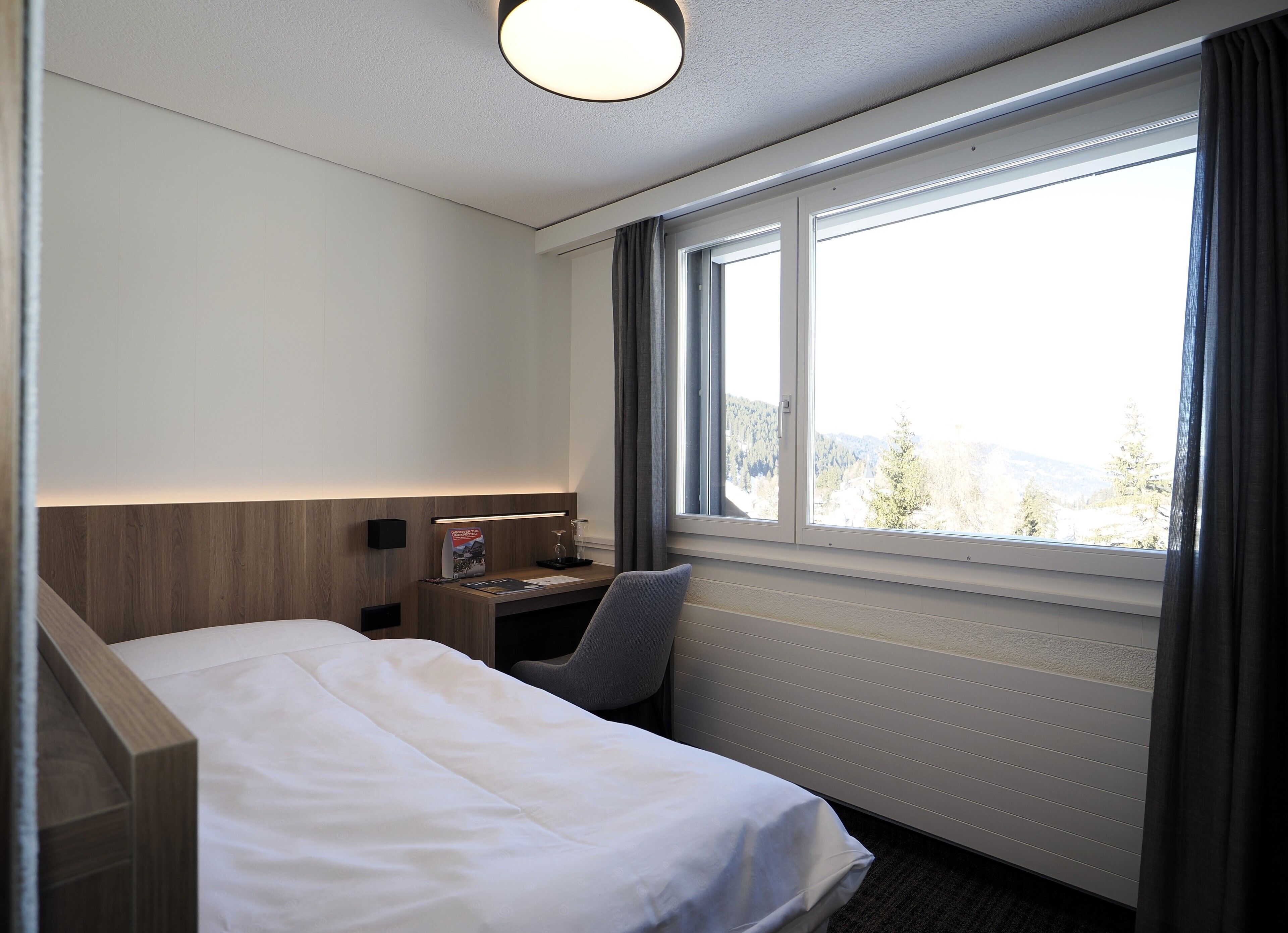 Una habitación de hotel con una cama, un escritorio con lámpara, una silla y un ventanal con vistas a árboles y montañas.