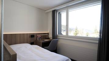 Una habitación de hotel con una cama, un escritorio con lámpara, una silla y un ventanal con vistas a árboles y montañas.