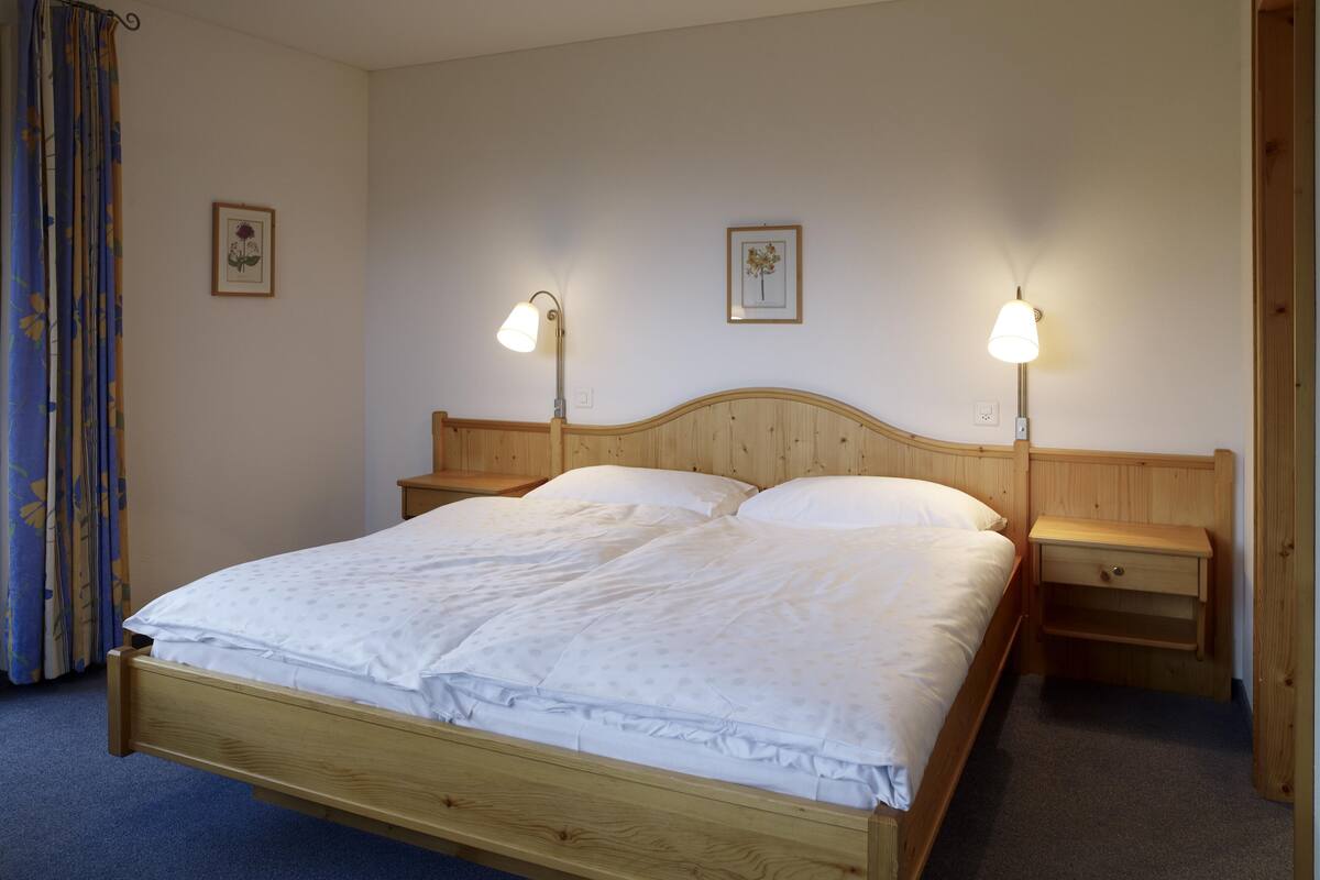 Un lit en bois, avec deux tables de chevet, chacune surmontée d’une lampe. Deux tableaux encadrés sont accrochés au mur.