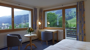 A hotel room with a bed, two chairs, a small table with a vase of flowers, and large windows offering a view of a green landscape. - Hotel Alpina Parpan (Churwalden)