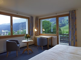 Habitación de hotel con cama, dos sillas, una mesita con un jarrón de flores y ventanas amplias que ofrecen vistas a un paisaje verde.