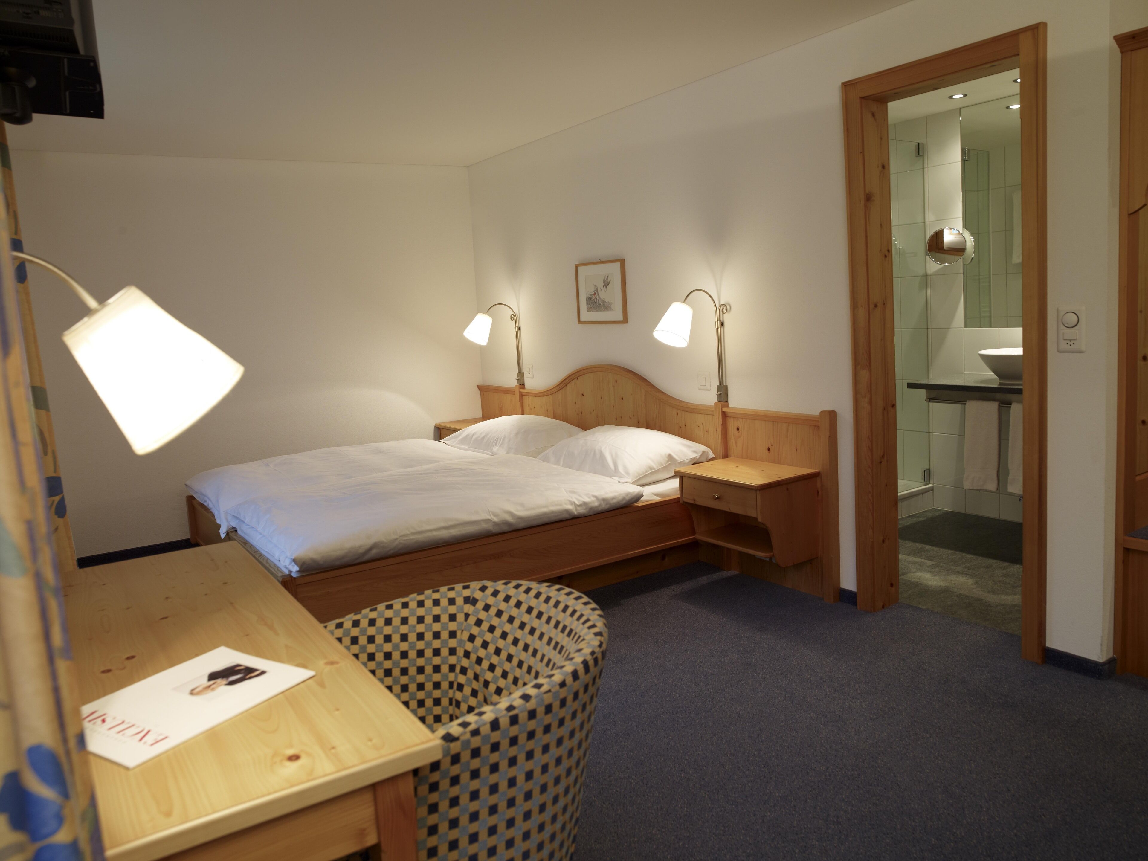 A bedroom with a wooden bed, two bedside lamps, a wooden desk, and a checkered chair.