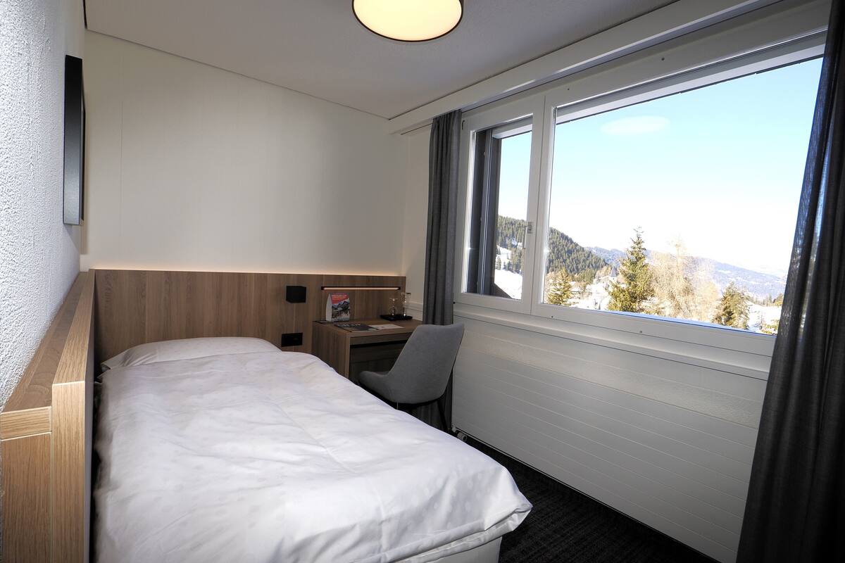 Une chambre d’hôtel équipée d’un lit, d’un bureau, d’une chaise et d’une fenêtre donnant sur les montagnes.