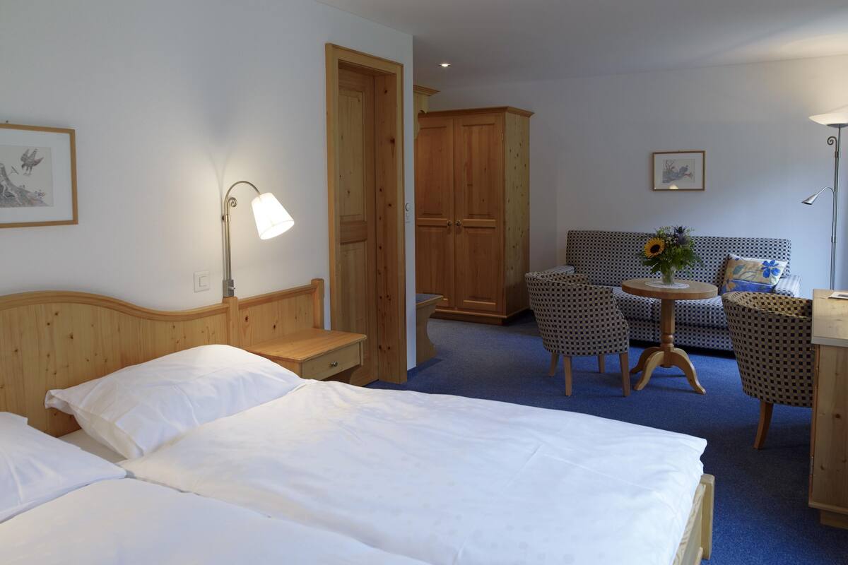 Une chambre d’hôtel avec deux lits, une armoire en bois, un bureau avec une lampe, un canapé à motifs et une petite table ronde sur laquelle est posé un vase.