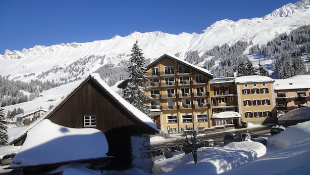 A snow-covered mountain resort with multi-story buildings, including a wooden chalet and a yellow building with balconies. - Hotel Alpina Parpan (Churwalden)