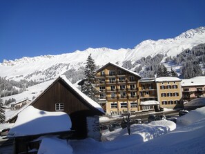 A snow-covered mountain resort with multi-story buildings, including a wooden chalet and a yellow building with balconies. - Hotel Alpina Parpan (Churwalden)