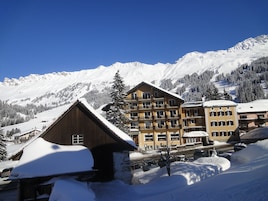 Un complejo turístico en la montaña cubierto de nieve, con edificios de varias plantas, incluyendo un chalet de madera y un edificio amarillo con balcones.