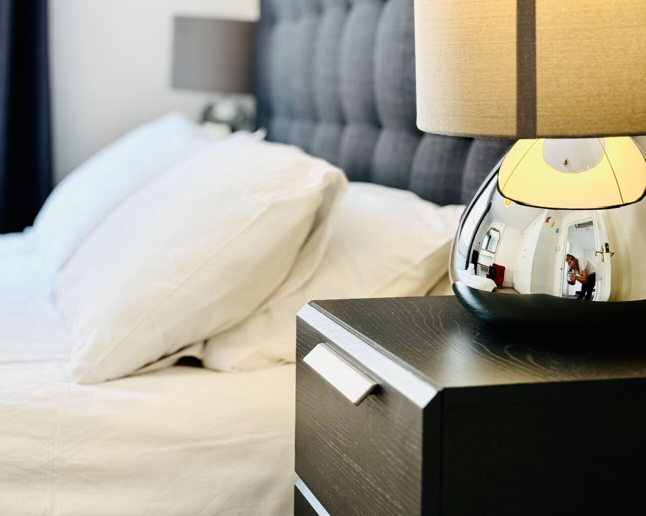 A neatly made bed with white bedding, a bedside lamp, and a nightstand with a reflective surface.