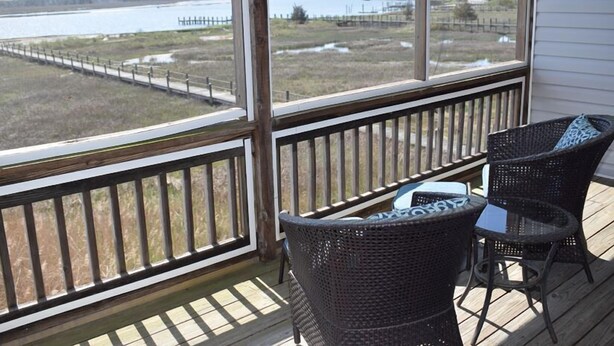 A wooden deck with wicker chairs and a table, overlooking a lake and marshland.