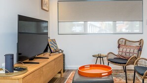 Un salon moderne doté d’un téléviseur à écran plat, d’une console en bois, de fauteuils en osier et d’une grande fenêtre avec un store enrouleur.
