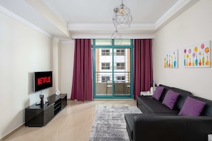A modern living room with a flat-screen TV displaying 'NETFLIX', a black sofa with purple cushions, a glass coffee table, and a geometric pendant light. - Delighful 1BR with coast view (Dubai)