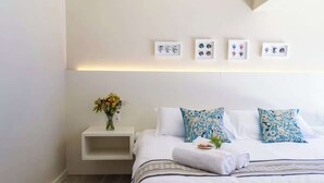 Une chambre d’hôtel moderne avec un lit soigneusement fait, une petite table de chevet ornée d’un vase de fleurs et trois tableaux encadrés accrochés au mur au-dessus du lit.