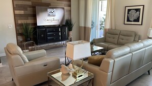 A living room with a flat-screen TV mounted on a wooden wall, a beige sofa, a glass coffee table, and a side table with a lamp. - Luxury/Modern 5Bed and 4bath Private Pool and Free Resort 10min da Disney 11ppl (Kissimmee)