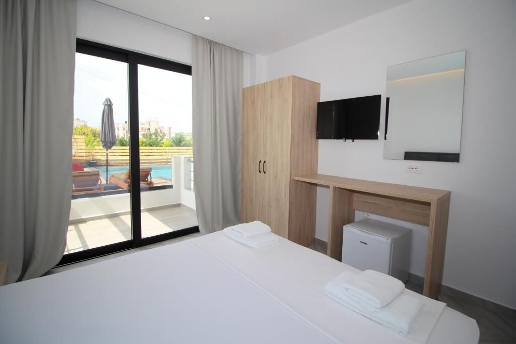 A hotel room with a bed, a desk with a computer, a TV, and a large window with a view of a pool and outdoor seating.