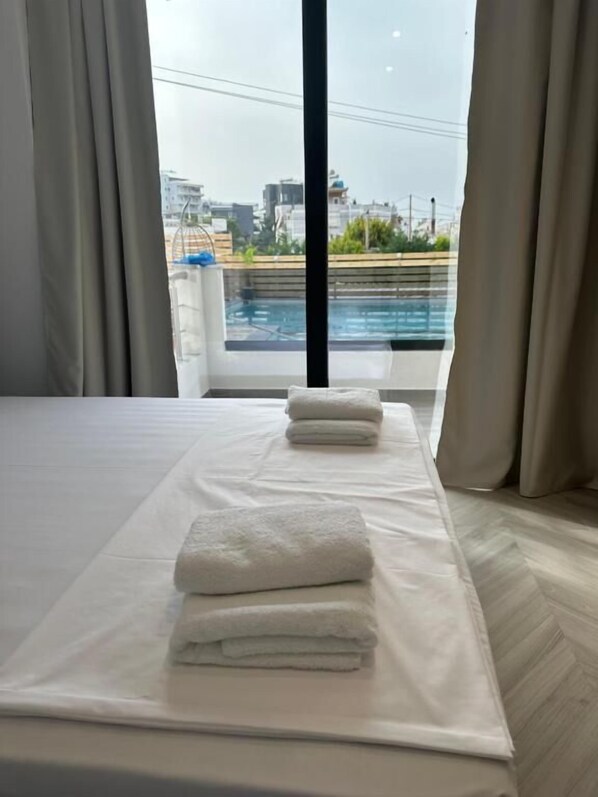 Une chambre d’hôtel avec un lit, deux serviettes et une vue sur une piscine et le paysage urbain, le tout visible à travers de grandes fenêtres.