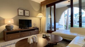 Habitación de hotel moderna con sofá, televisión sobre una consola de madera, lámpara y vistas a palmeras a través de amplias ventanas.