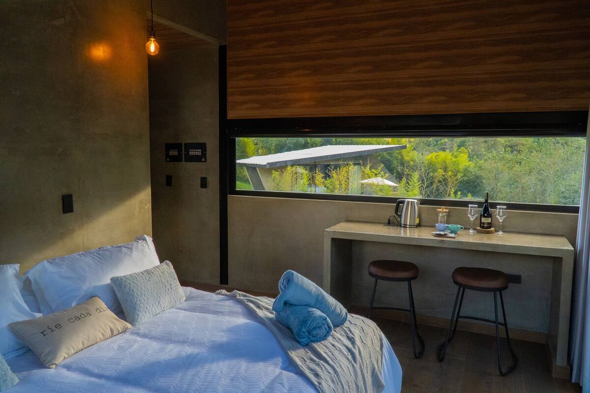 A modern bedroom with a large window, a bed with pillows, a desk with stools, and a view of greenery outside.
