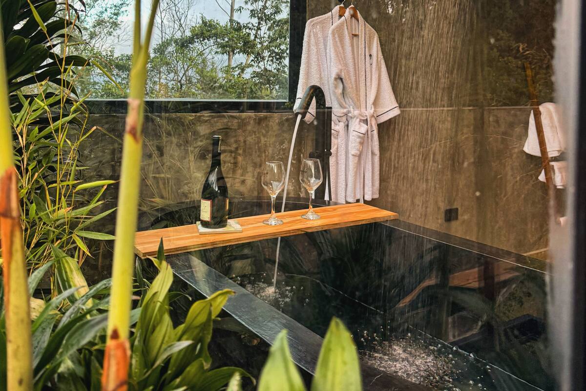 A modern bathroom with a rain shower, a wooden shelf with a bottle and glasses, and a towel rack.