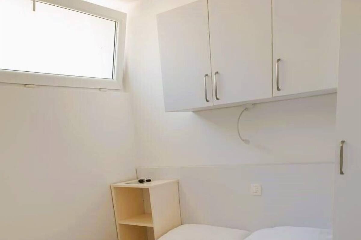 A single-bed room with a white bed, a small wooden nightstand, and a white wardrobe with two doors.