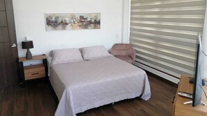 Ein Schlafzimmer mit Bett, Nachttisch, Lampe, Stuhl, Fernseher und Fenster mit Jalousien.