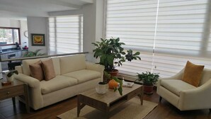 Ein modernes Wohnzimmer mit einem beigen Sofa, einem Holztisch und zwei Sesseln. Auf dem Tisch stehen Blumentöpfe und im Hintergrund ist eine Küchenzeile erkennbar.