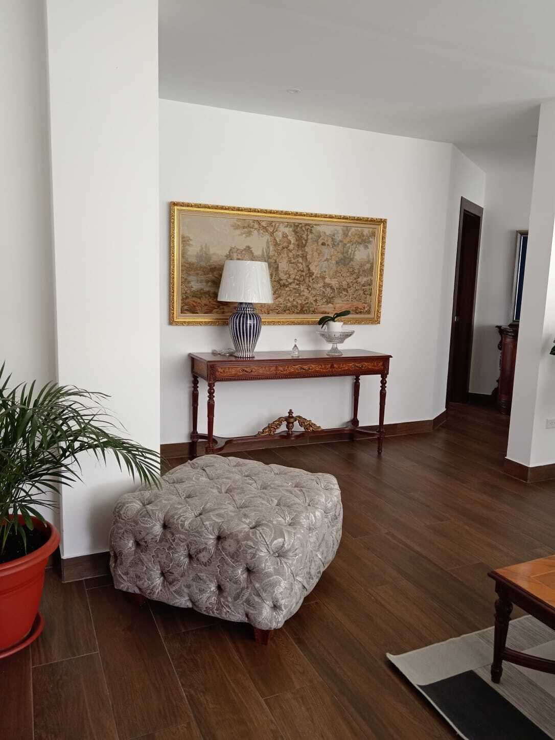 A living room with a large painting, a table with a lamp, a potted plant, and a tufted ottoman.