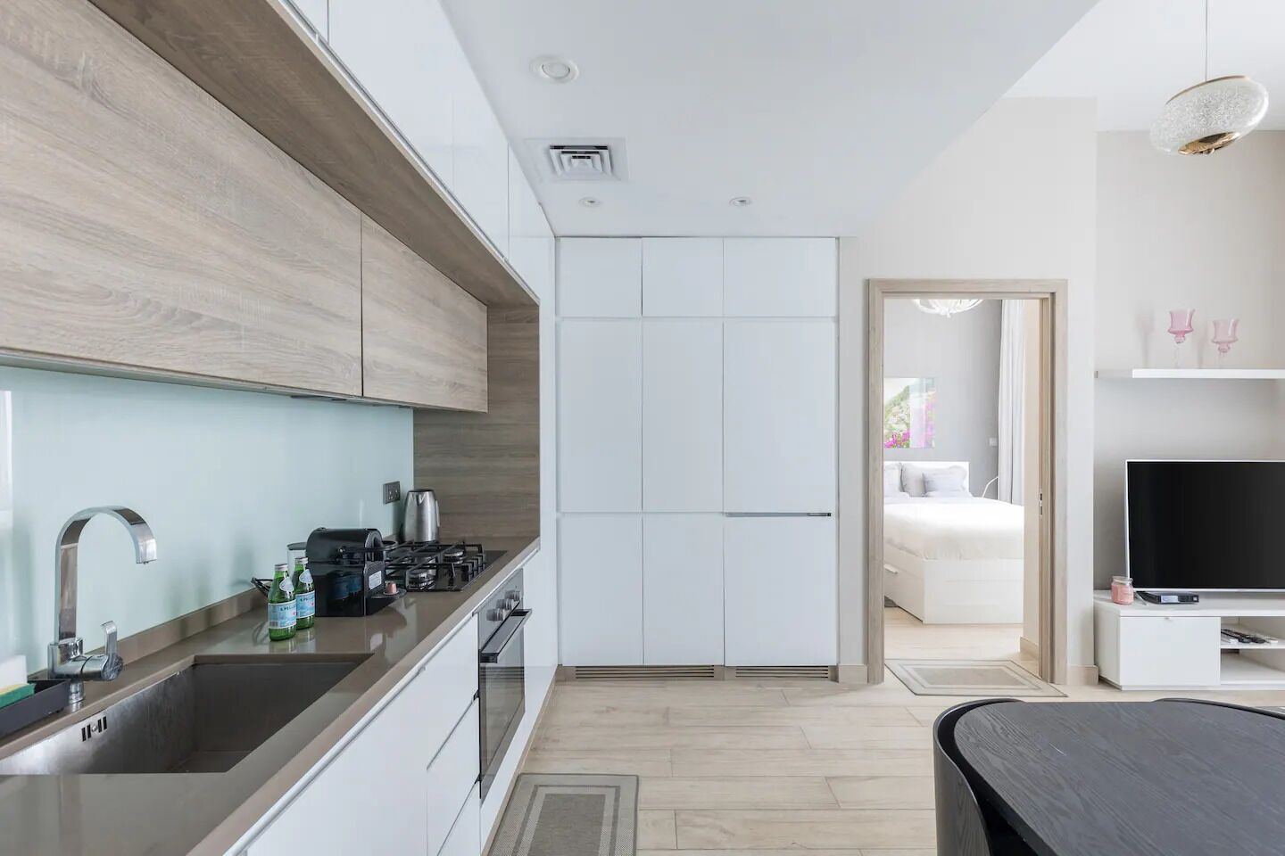 A modern kitchen with a sink, stove, and cabinets, leading to a bedroom with a bed and a TV.