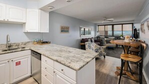 A modern kitchen with white cabinets, a granite countertop, and a built-in dishwasher. There is a dining area with a round table and bar stools. - Escape to paradise! 1BR Beachfront Condo in Destin (Destin)
