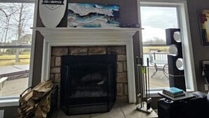 A living room with a stone fireplace, large windows, and artwork on the walls. - Avon Lake, Avon golf course home, 1,000 s.f. open outdoor deck on course lake (Avon)