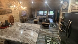 Interior acogedor de una cabaña con una isla grande de mármol en la cocina, una chimenea de piedra, paredes de madera y vistas a través de las ventanas.