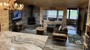 Interior acogedor de una cabaña con chimenea de piedra, techo de madera y un televisor en la pared.
