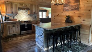 Una cocina con armarios de madera, encimera de mármol, una isla central con taburetes de bar, una araña y decoración rústica en la pared.