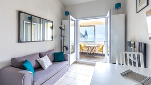 A modern room with a sofa, a dining table, a TV, and a large window offering a view of the sea.