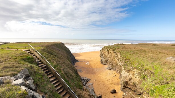 A coastal landscape with a staircase leading down to a beach, surrounded by grassy terrain and cliffs.