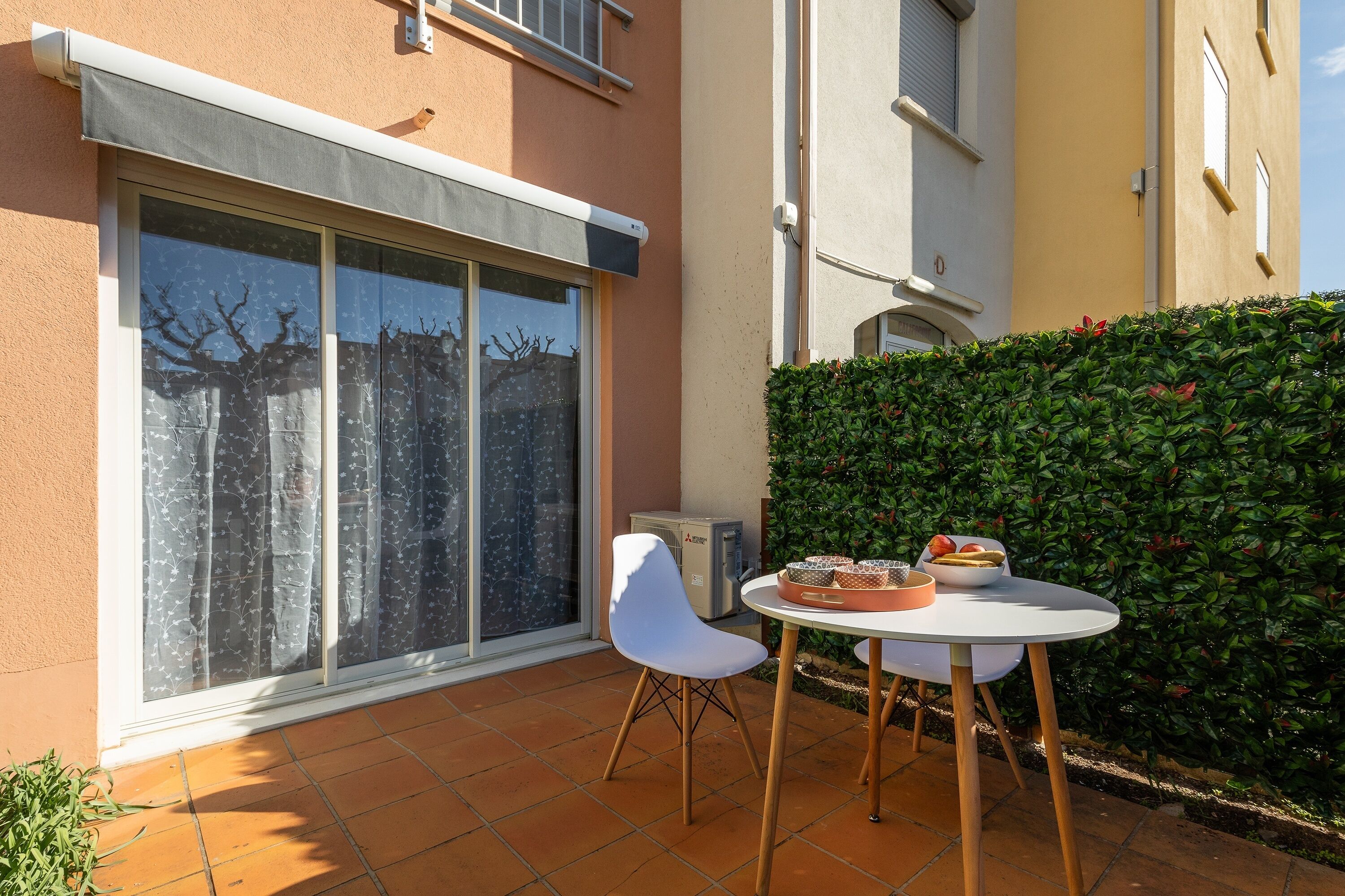 Un patio con una mesa y sillas blancas, una mesita con fruta y un amplio ventanal corredero.