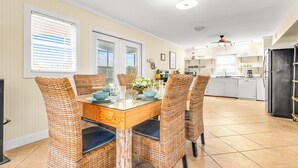 A dining area with a glass-top table surrounded by wicker chairs, a kitchen in the background, and a ceiling fan.