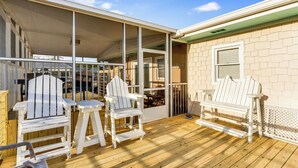 A covered deck with white Adirondack chairs and a bench, wooden flooring, and a brick building with a window.