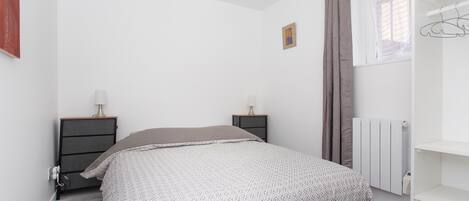 A bedroom with a bed, two bedside tables, a radiator, and a window with curtains.