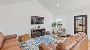 A spacious living room with a high ceiling, a large TV mounted on the wall, a wooden entertainment center, a brown leather sofa, a grey armchair, and a patterned rug. - BLUEBERRY COTTAGE is located in Mena.  WIFI, remodeled , Lockable bike storage ! (Mena)