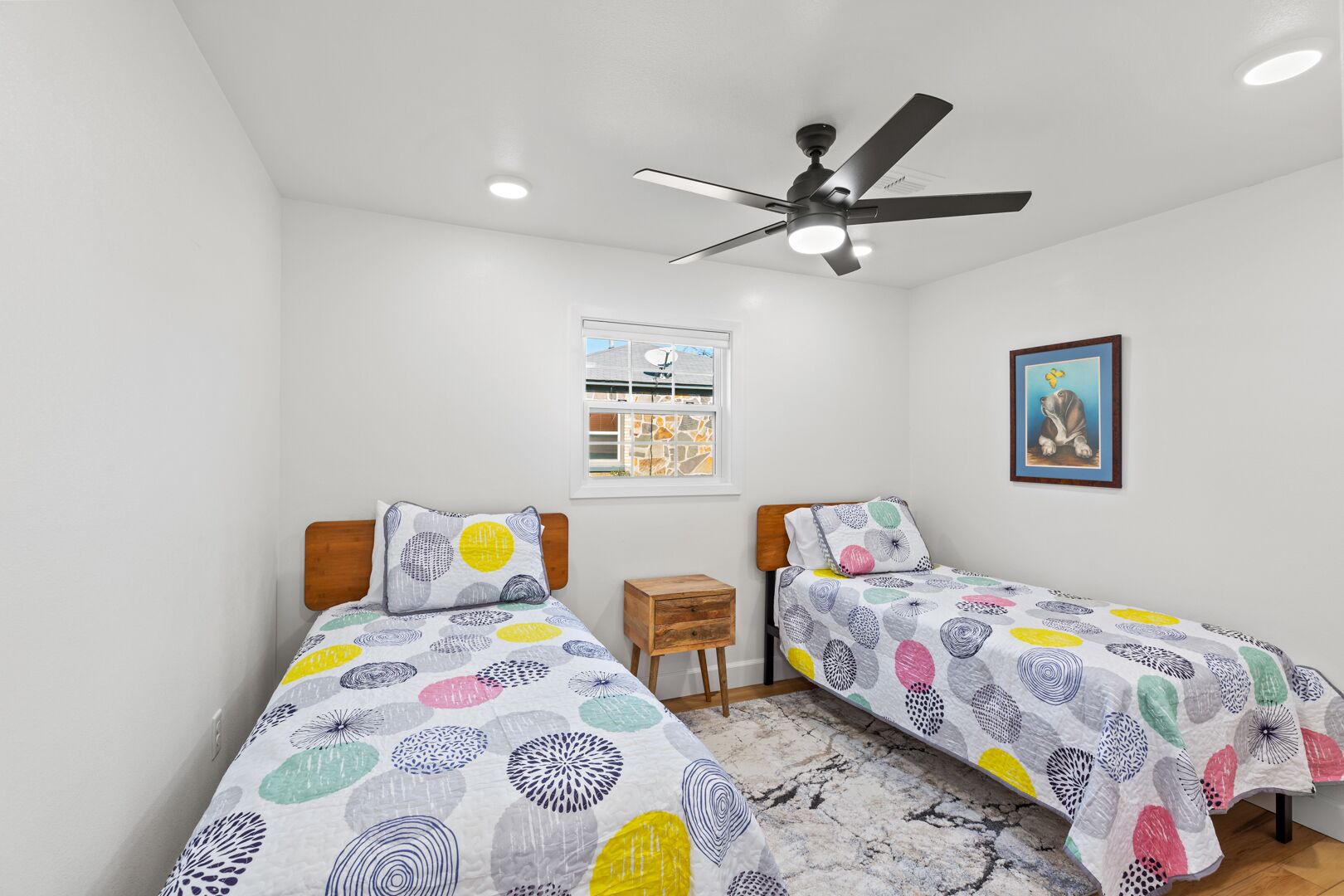 A bedroom with two single beds, a ceiling fan, a small wooden nightstand, a framed picture, and a window with a view of buildings.