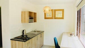 A compact kitchen with wooden cabinets, a black countertop, a stainless steel sink, and a kettle. There are two framed pictures on the wall and a window with bars. - Splendid 1-bed Apartment in Sandton (Johannesburg)