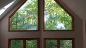 Ein Zimmer mit schrägem Dach, Deckenventilator und vier Fenstern mit Blick auf einen Wald.