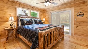 Ein Schlafzimmer einer Holzhütte mit einem Bett, zwei Nachttischen, einem Deckenventilator und einem Fenster mit Jalousien.